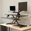 Front view of a black electric standing desk converter fully raised, showing the laptop tray, monitor arm, main work surface, and keyboard tray with a keyboard and mouse.